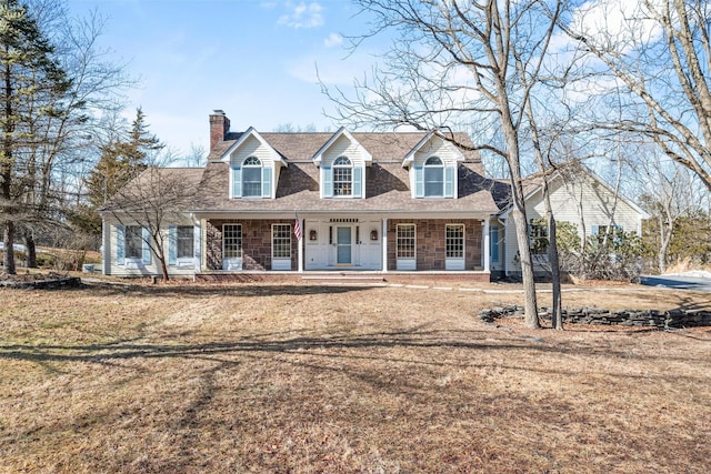 cape cod house featuring covered porch, stone siding, roof with shingles, a chimney, and a front yard