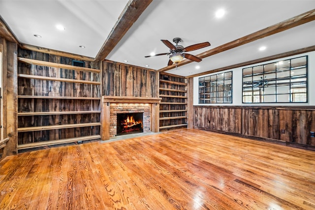 unfurnished living room with wooden walls, wood finished floors, a brick fireplace, built in shelves, and beam ceiling