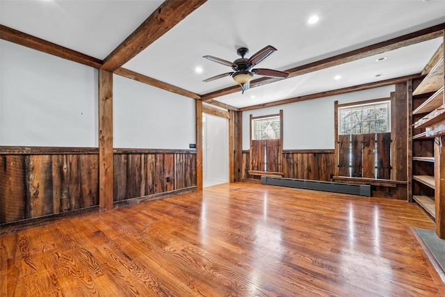 unfurnished room with wood finished floors, a ceiling fan, baseboard heating, wainscoting, and beam ceiling