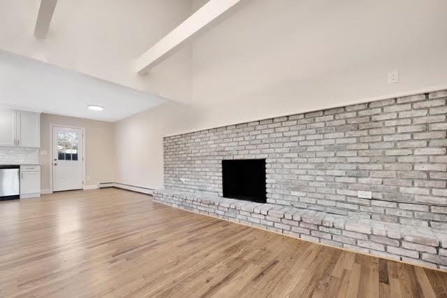 unfurnished living room with baseboard heating, lofted ceiling with beams, a brick fireplace, and light wood-type flooring