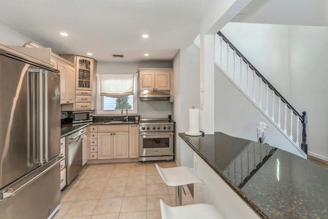 kitchen with sink, light tile patterned floors, high end appliances, light brown cabinetry, and dark stone counters