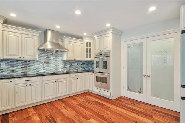 kitchen featuring light wood-type flooring, wall chimney exhaust hood, black electric cooktop, and double oven