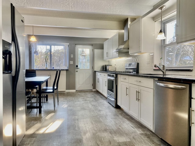 kitchen featuring wall chimney exhaust hood, decorative light fixtures, light wood-type flooring, appliances with stainless steel finishes, and white cabinets