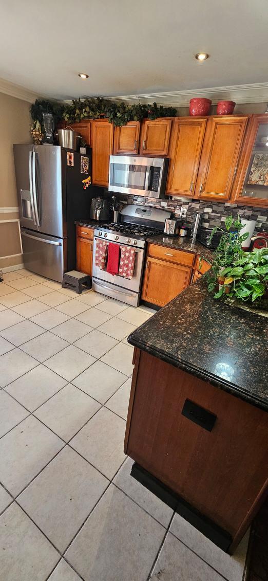 kitchen featuring light tile patterned floors, dark stone countertops, stainless steel appliances, ornamental molding, and decorative backsplash