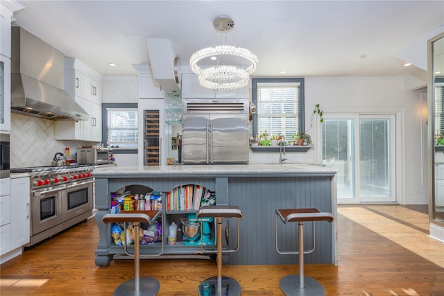 kitchen with wall chimney range hood, hardwood / wood-style floors, hanging light fixtures, high quality appliances, and white cabinets