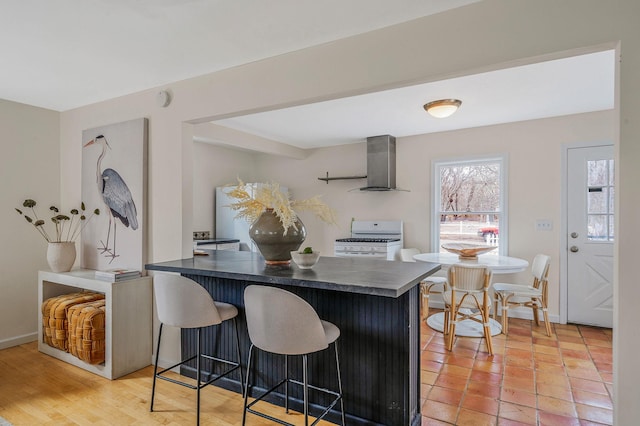 kitchen featuring white range with gas stovetop, a kitchen bar, kitchen peninsula, wall chimney exhaust hood, and light wood-type flooring