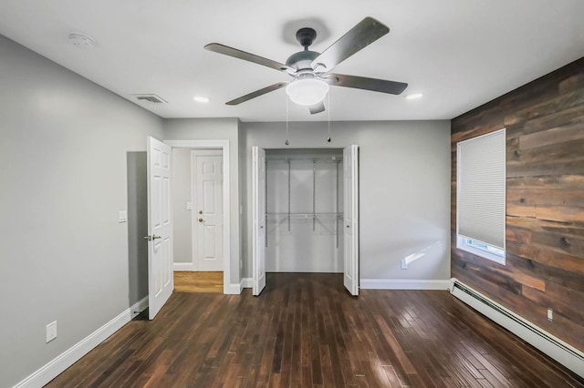 unfurnished bedroom featuring ceiling fan, a baseboard heating unit, wooden walls, dark hardwood / wood-style flooring, and a closet