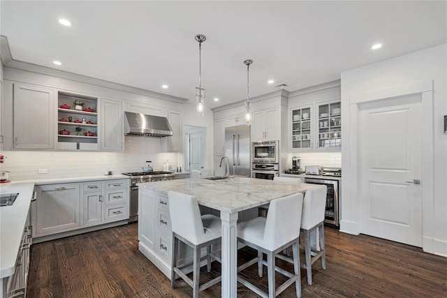 kitchen featuring wall chimney exhaust hood, sink, built in appliances, an island with sink, and white cabinets