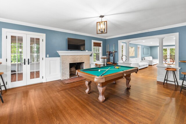 rec room featuring french doors, crown molding, a brick fireplace, hardwood / wood-style floors, and decorative columns