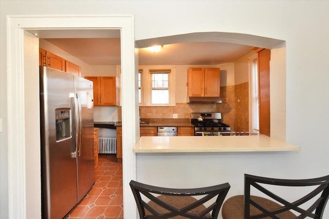 kitchen with a breakfast bar area, dark tile patterned floors, appliances with stainless steel finishes, radiator heating unit, and decorative backsplash