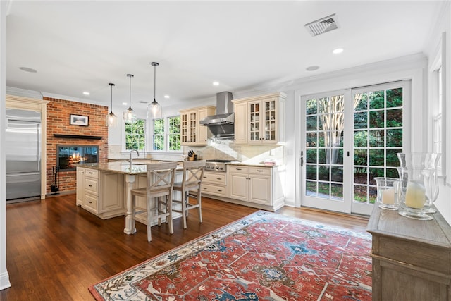 kitchen with decorative light fixtures, light stone counters, stainless steel appliances, a center island with sink, and wall chimney exhaust hood