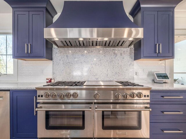 155 Pinewood Rd, Cutchogue, NY 11935 - kitchen with light stone countertops, blue cabinetry, range with two ovens, range hood, and backsplash