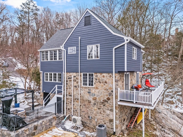 snow covered property with a deck, stone siding, roof with shingles, and central air condition unit