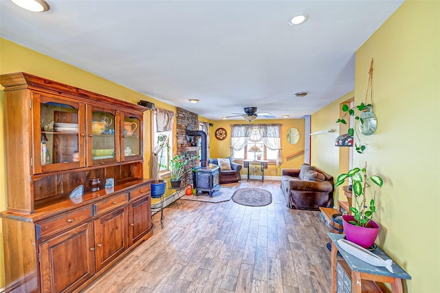 living room featuring ceiling fan, light wood-style flooring, baseboard heating, and a wood stove