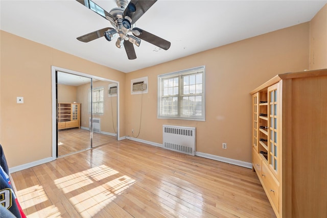 unfurnished bedroom featuring a wall unit AC, light wood finished floors, radiator heating unit, a ceiling fan, and baseboards