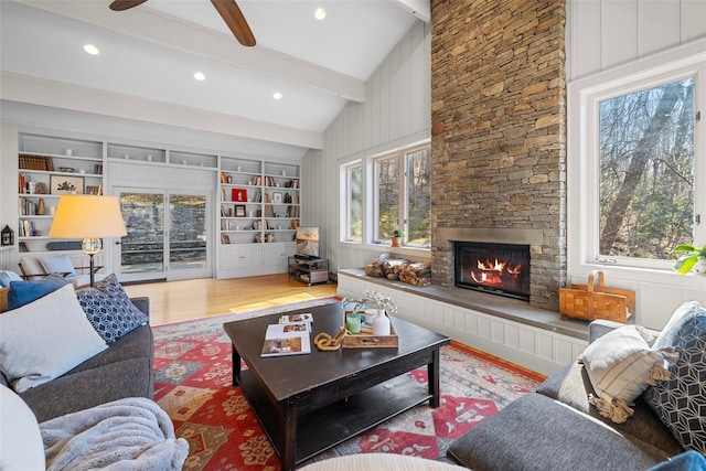 living area featuring built in features, wood finished floors, lofted ceiling with beams, ceiling fan, and a stone fireplace