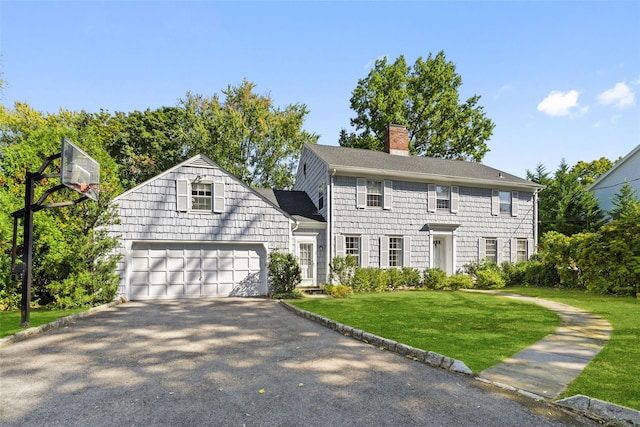 colonial house with a chimney, aphalt driveway, roof with shingles, an attached garage, and a front yard