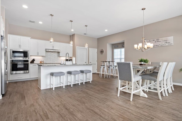 kitchen with a breakfast bar area, appliances with stainless steel finishes, white cabinets, a center island with sink, and decorative light fixtures