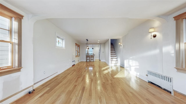 unfurnished living room featuring arched walkways, baseboards, radiator, stairway, and light wood-type flooring