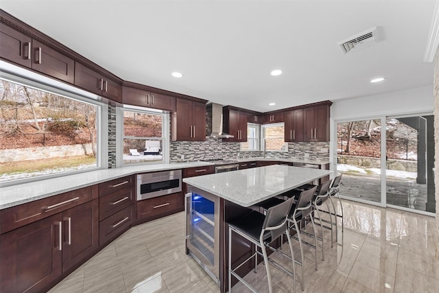 kitchen featuring a breakfast bar, a center island, stainless steel microwave, light stone countertops, and wall chimney range hood