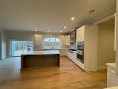 kitchen with double oven, white cabinets, a center island, light hardwood / wood-style floors, and wall chimney range hood