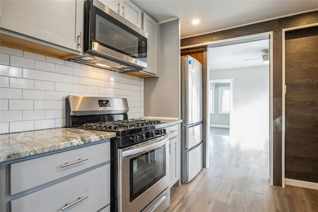 kitchen with gray cabinets, backsplash, light stone counters, stainless steel appliances, and light wood-type flooring