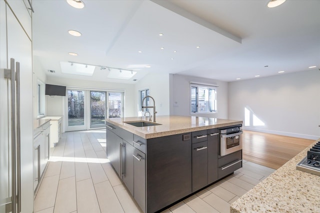 kitchen with sink, stainless steel oven, a center island with sink, light stone countertops, and light hardwood / wood-style floors