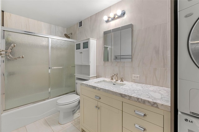 full bathroom with stacked washing maching and dryer, tile walls, tile patterned flooring, enclosed tub / shower combo, and toilet