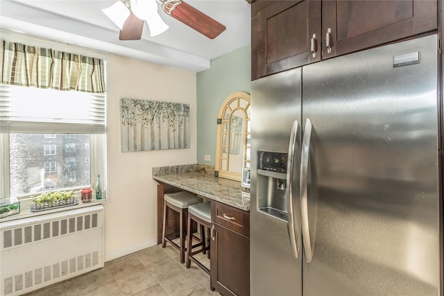 kitchen with radiator, stainless steel fridge, dark brown cabinetry, ceiling fan, and light stone countertops