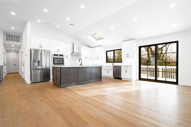 kitchen with appliances with stainless steel finishes, light hardwood / wood-style floors, white cabinets, a center island with sink, and wall chimney exhaust hood