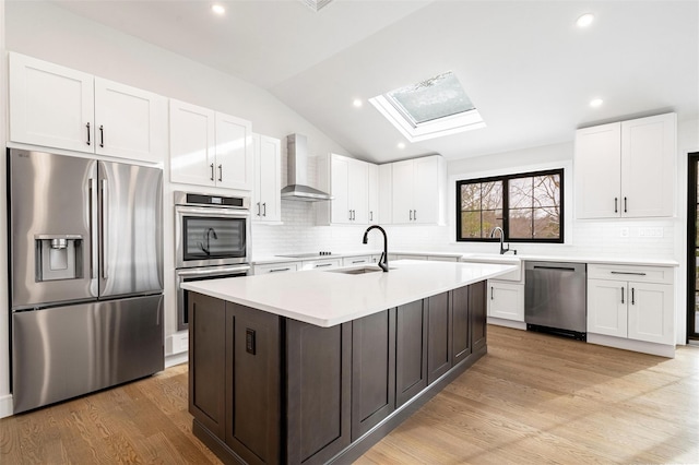 kitchen featuring wall chimney range hood, sink, appliances with stainless steel finishes, a kitchen island with sink, and white cabinets