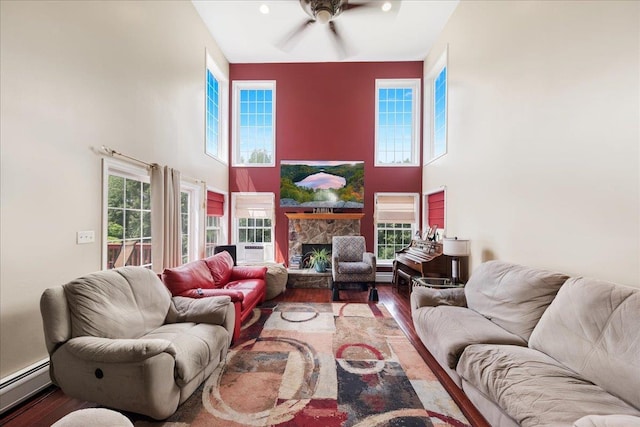 living room with ceiling fan, baseboard heating, hardwood / wood-style floors, a high ceiling, and a stone fireplace