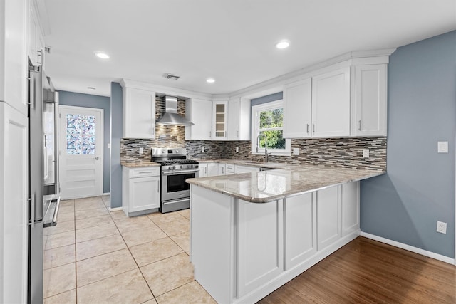 kitchen with wall chimney exhaust hood, white cabinetry, light stone counters, kitchen peninsula, and stainless steel appliances