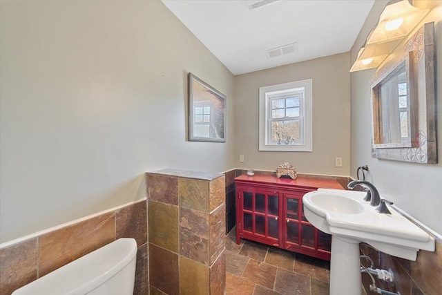 half bath with stone tile floors, visible vents, toilet, a wainscoted wall, and tile walls