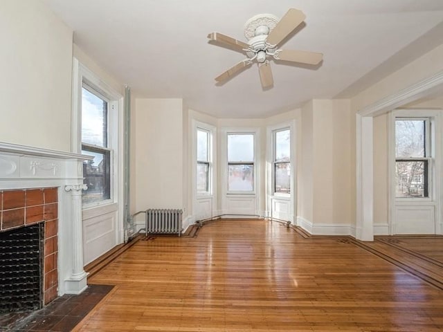 unfurnished living room with a tiled fireplace, ceiling fan, wood-type flooring, and radiator