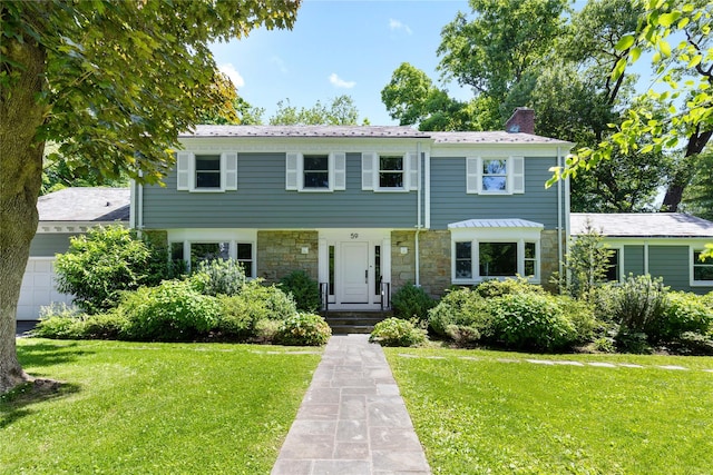 colonial-style house featuring stone siding, a front yard, an attached garage, and a chimney