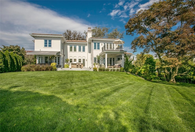 back of property with a lawn, a balcony, a tile roof, a chimney, and stucco siding