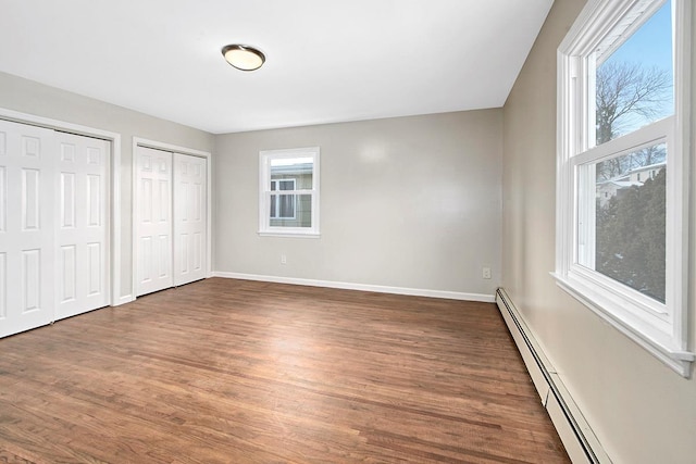 unfurnished bedroom with multiple windows, a baseboard heating unit, two closets, and dark wood-type flooring