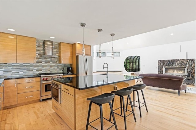 kitchen with pendant lighting, wall chimney range hood, a breakfast bar, appliances with stainless steel finishes, and a kitchen island with sink