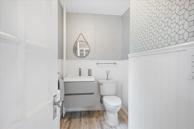 half bath featuring toilet, a wainscoted wall, wood finished floors, vanity, and wallpapered walls