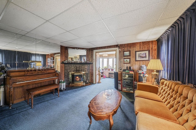 living room featuring wood walls, carpet flooring, a stone fireplace, and a drop ceiling