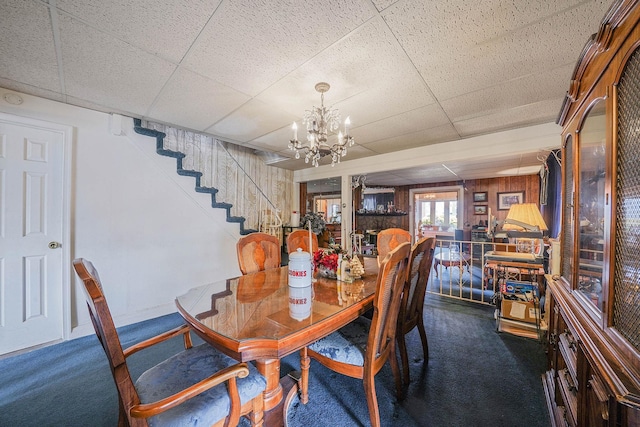 dining area with wood walls, a chandelier, a drop ceiling, and dark colored carpet