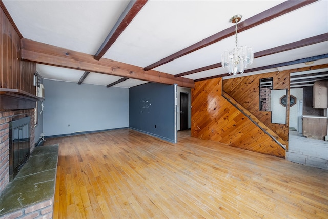 unfurnished living room featuring wood walls, beam ceiling, a brick fireplace, an inviting chandelier, and light hardwood / wood-style flooring