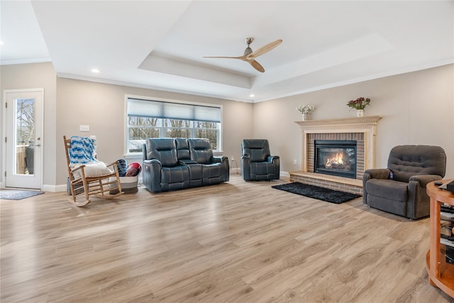 living room featuring light hardwood / wood-style flooring, ceiling fan, a tray ceiling, a fireplace, and ornamental molding