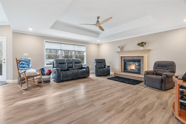 living room featuring ceiling fan, a raised ceiling, a brick fireplace, and light wood-type flooring