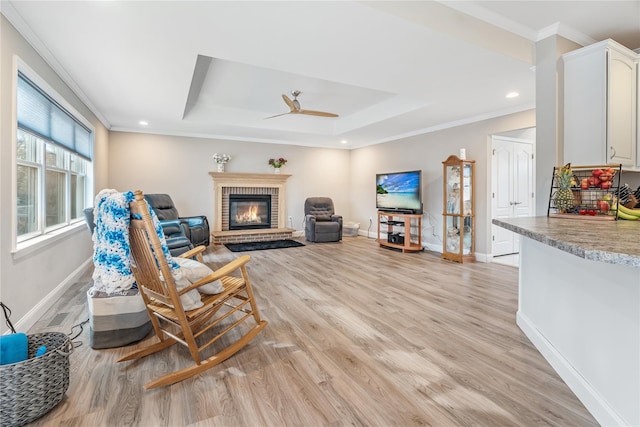 living room with a brick fireplace, light hardwood / wood-style flooring, ornamental molding, a tray ceiling, and ceiling fan