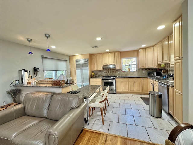 kitchen featuring dark stone countertops, stainless steel appliances, tasteful backsplash, decorative light fixtures, and light brown cabinets