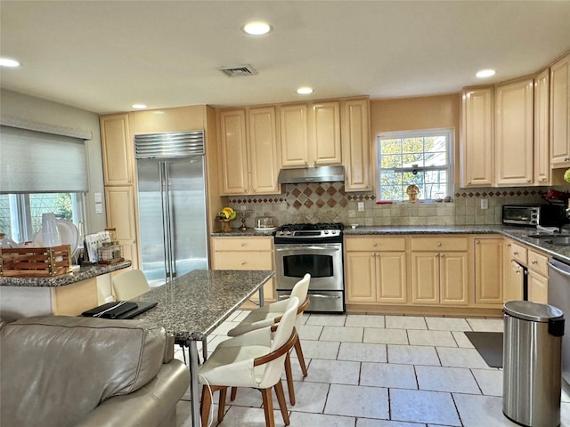 kitchen with appliances with stainless steel finishes, dark stone countertops, backsplash, and light brown cabinets