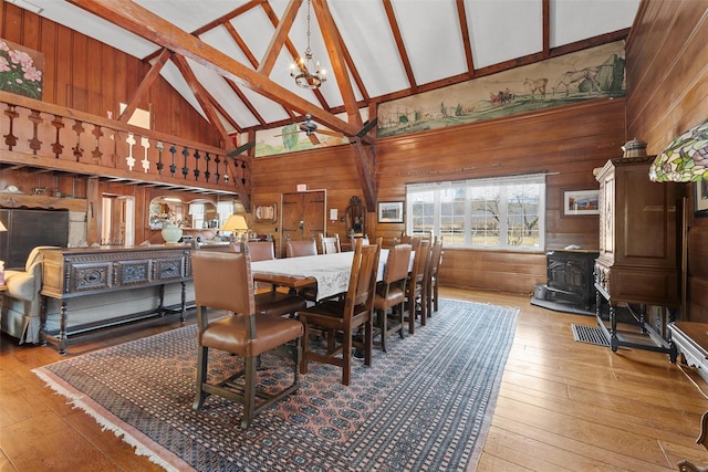 dining space with light wood-style flooring, a wood stove, wooden walls, high vaulted ceiling, and ceiling fan with notable chandelier