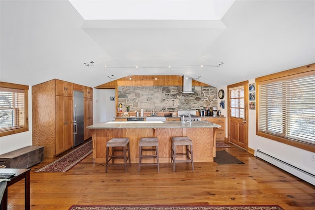 kitchen with light countertops, baseboard heating, brown cabinetry, wall chimney range hood, and stainless steel built in refrigerator